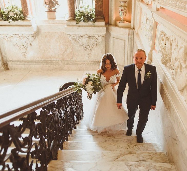 Bride wearing Martina Liana dress and her father walking up the stairs styled with white foliage decor