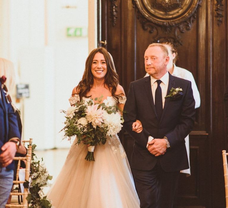 Bride wearing Martina Liana dress and veil walking down the aisle clutching white floral bouquet