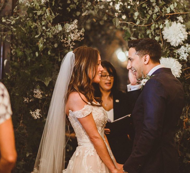 Bride wearing Martina Liana off the shoulder dress with veil holds hands with the groom at wedding ceremony