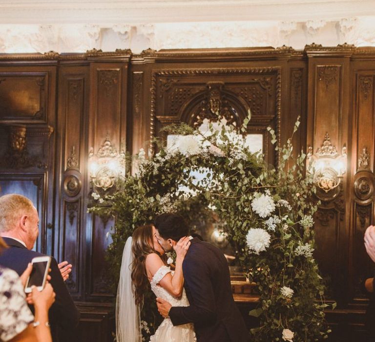 Bride and groom kiss with white foliage archway and candles scattered with petals