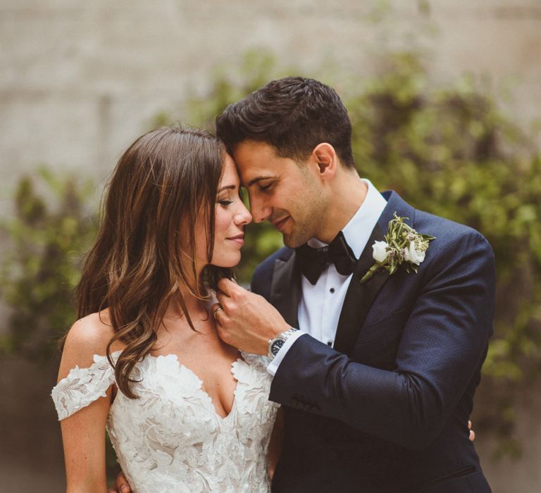 Bride wearing laced Martina Liana dress and groom wearing black tie suit with white floral buttonhole