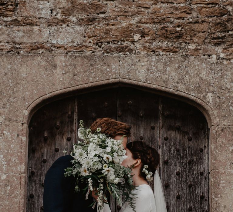 Bride in Homemade Wedding Dress with Long Sleeves and Groom in Navy Reiss Suit Kissing Behind a White and Green Bouquet