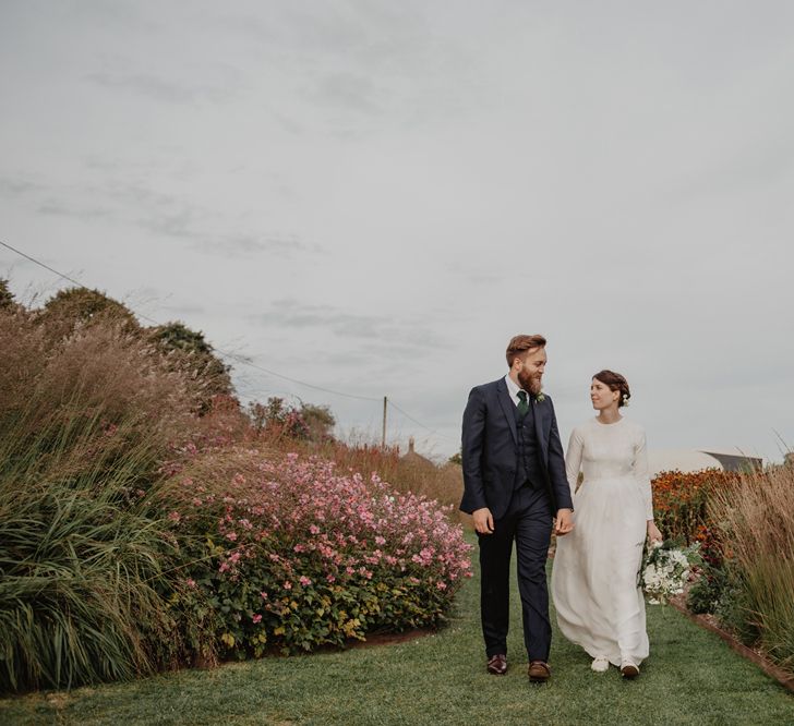 Bride in Homemade Wedding Dress with Long Sleeves and Groom in Navy Reiss Suit Holding Hands