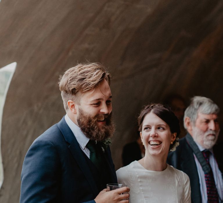 Bride in Homemade Wedding Dress with Long Sleeves and Groom in Navy Reiss Suit Laughing