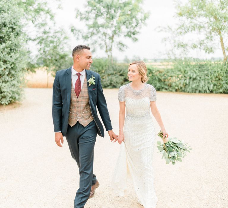 Bride in Beaded Wedding Dress and Groom in Dark Suit &amp; Check Waistcoat Holding Hands