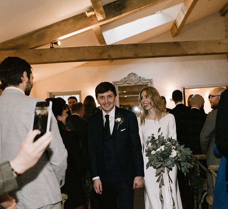 Bride in Long Sleeved Andrea Hawkes Dress with Bateau Neck and Low V-Back with Buttons | Groom in Navy Three-Piece Suit from Suit Supply with Green Tie | Bridal Bouquet with Eucalyptus, White Roses, White and Blue Thistles and Ferns | Emerald Green Bridesmaid Dress for a Winter Wedding at Middleton Lodge | Georgina Harrison Photography