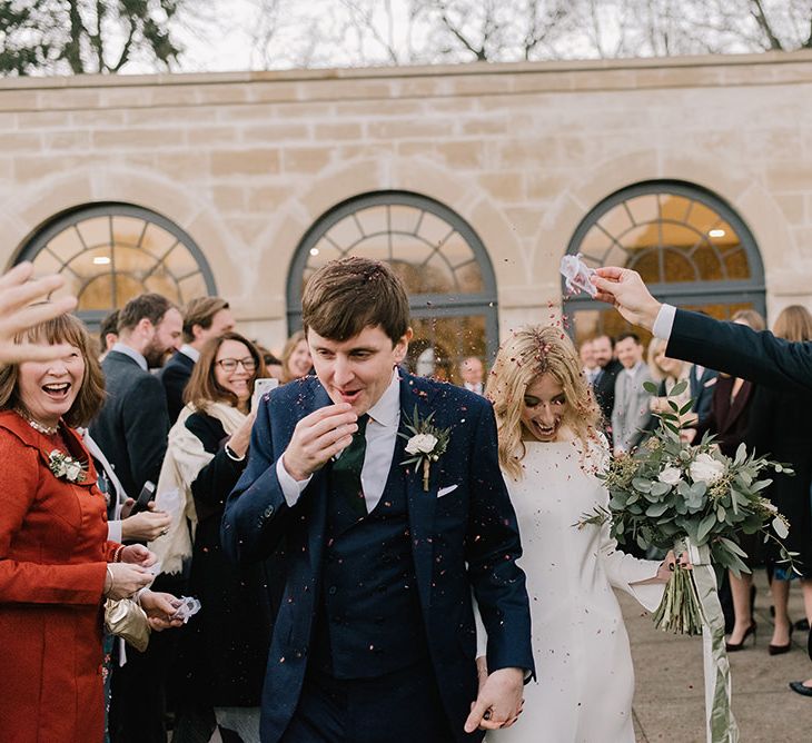 Bride in Long Sleeved Andrea Hawkes Dress with Bateau Neck and Low V-Back with Buttons | Groom in Navy Three-Piece Suit from Suit Supply with Green Tie | Bridal Bouquet with Eucalyptus, White Roses, White and Blue Thistles and Ferns | Emerald Green Bridesmaid Dress for a Winter Wedding at Middleton Lodge | Georgina Harrison Photography