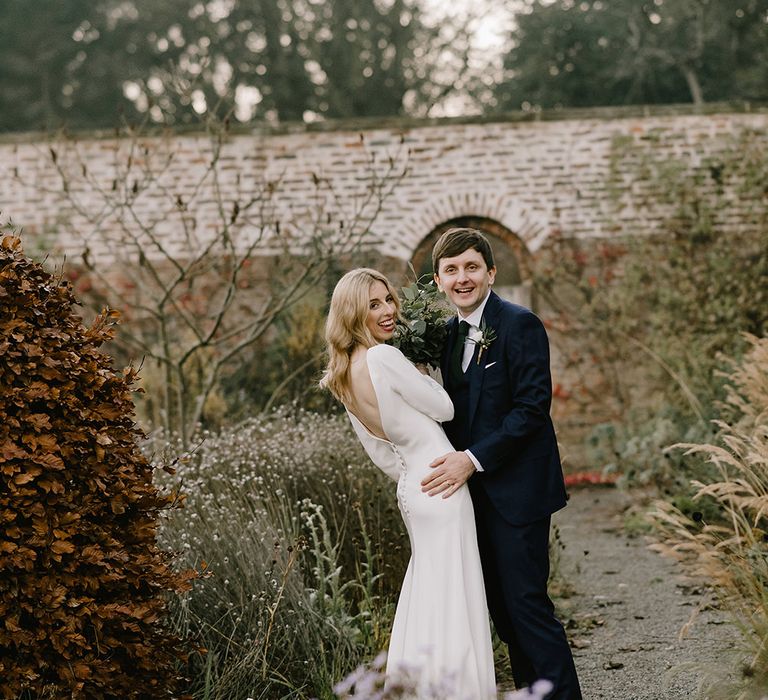 Bride in Long Sleeved Andrea Hawkes Dress with Bateau Neck and Low V-Back with Buttons | Groom in Navy Three-Piece Suit from Suit Supply with Green Tie | Bridal Bouquet with Eucalyptus, White Roses, White and Blue Thistles and Ferns | Emerald Green Bridesmaid Dress for a Winter Wedding at Middleton Lodge | Georgina Harrison Photography