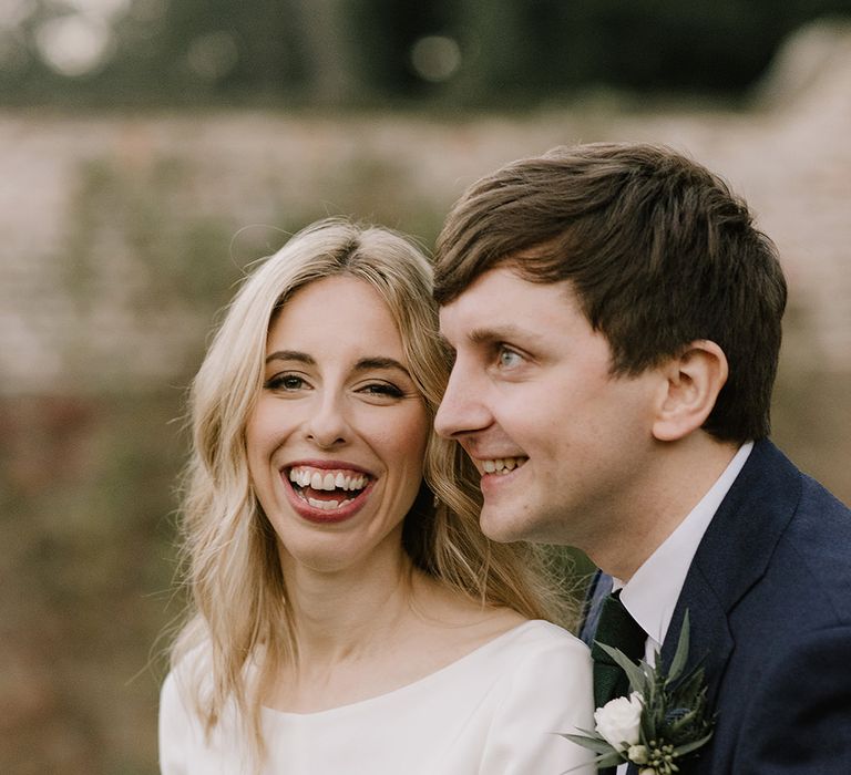 Bride in Long Sleeved Andrea Hawkes Dress with Bateau Neck and Low V-Back with Buttons | Groom in Navy Three-Piece Suit from Suit Supply with Green Tie | Bridal Bouquet with Eucalyptus, White Roses, White and Blue Thistles and Ferns | Emerald Green Bridesmaid Dress for a Winter Wedding at Middleton Lodge | Georgina Harrison Photography