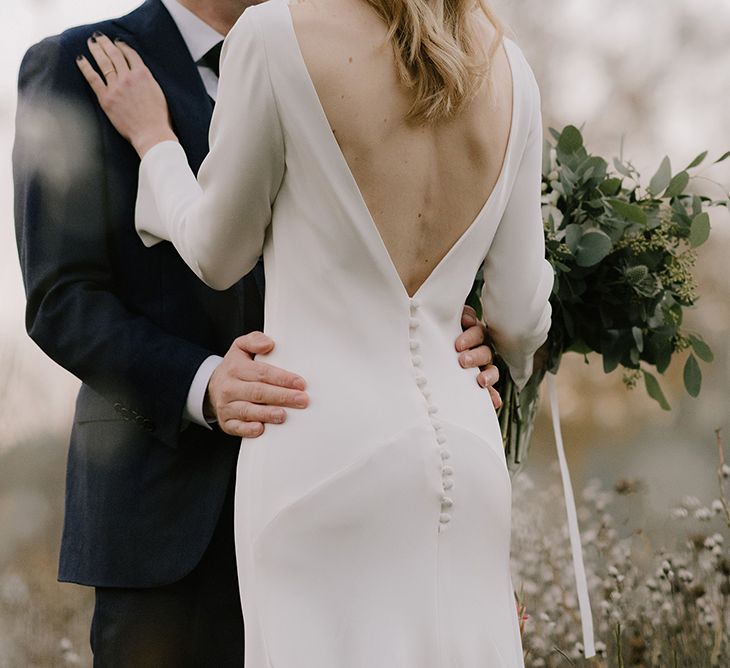 Bride in Long Sleeved Andrea Hawkes Dress with Bateau Neck and Low V-Back with Buttons | Groom in Navy Three-Piece Suit from Suit Supply with Green Tie | Bridal Bouquet with Eucalyptus, White Roses, White and Blue Thistles and Ferns | Emerald Green Bridesmaid Dress for a Winter Wedding at Middleton Lodge | Georgina Harrison Photography