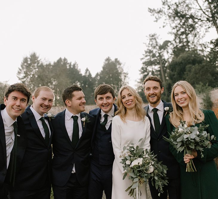 Bride in Long Sleeved Andrea Hawkes Dress with Bateau Neck and Low V-Back with Buttons | Groom in Navy Three-Piece Suit from Suit Supply with Green Tie | Bridesmaid in Forest Green Reformation Dress with Long Sleeves | Wedding Bouquets with Eucalyptus, White Roses, White and Blue Thistles and Ferns | Groomsmen in Navy Marks &amp; Spencer Suits with Green Ties | Emerald Green Bridesmaid Dress for a Winter Wedding at Middleton Lodge | Georgina Harrison Photography