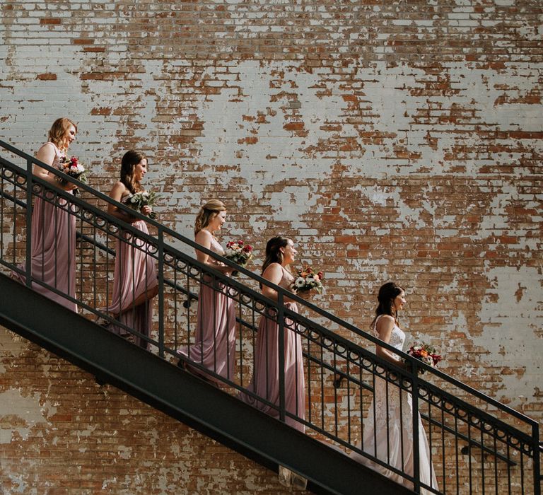 Bridal Party on Staircase at BRIK | Bride in Sleeveless High-Neck Stella York Gown with Lace Overlay and Gold Underskirt | Bridesmaids in Pink Dresses with Lace Bodice | Colourful Bouquets of Pink, Orange and White Flowers with Ferns, Eucalyptus and Greenery | Rifle Paper Co. Trainers for Informal Wedding in Fort Worth, Texas | Paul &amp; Nanda Photography