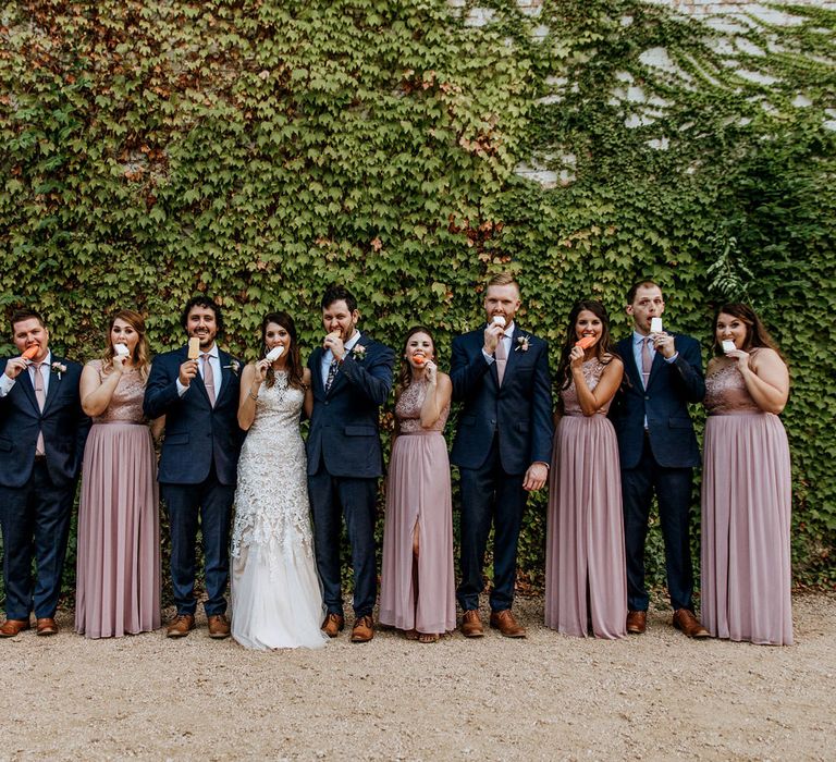 Bridal Party Eating Popsicles | Bride in Sleeveless High-Neck Stella York Gown with Lace Overlay and Gold Underskirt | Groom in Navy Suit and Floral Tie | Bridesmaids in Pink Dresses with Lace Bodice | Groomsmen in Navy Suits and Silk Blush Ties | Pink Buttonholes | Rifle Paper Co. Trainers for Informal Wedding in Fort Worth, Texas | Paul &amp; Nanda Photography