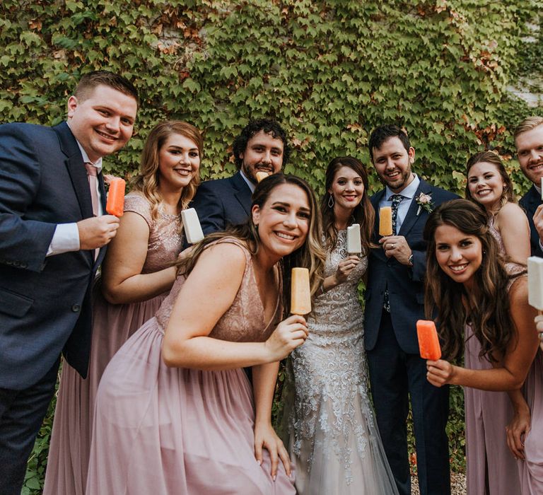 Bridal Party Eating Popsicles | Bride in Sleeveless High-Neck Stella York Gown with Lace Overlay and Gold Underskirt | Groom in Navy Suit and Floral Tie | Bridesmaids in Pink Dresses with Lace Bodice | Groomsmen in Navy Suits and Silk Blush Ties | Pink Buttonholes | Rifle Paper Co. Trainers for Informal Wedding in Fort Worth, Texas | Paul &amp; Nanda Photography