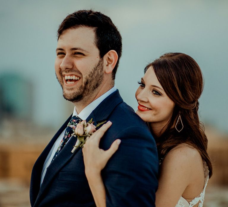 Bride in Sleeveless High-Neck Stella York Gown with Lace Overlay and Gold Underskirt | Half Up Half Down Wedding Hair | Bride Wearing Red Lipstick | Groom in Navy Suit and Floral Tie | Pink Buttonhole | Colourful Bridal Bouquet of Pink, Orange and White Flowers with Ferns, Eucalyptus and Greenery | Rifle Paper Co. Trainers for Informal Wedding in Fort Worth, Texas | Paul &amp; Nanda Photography