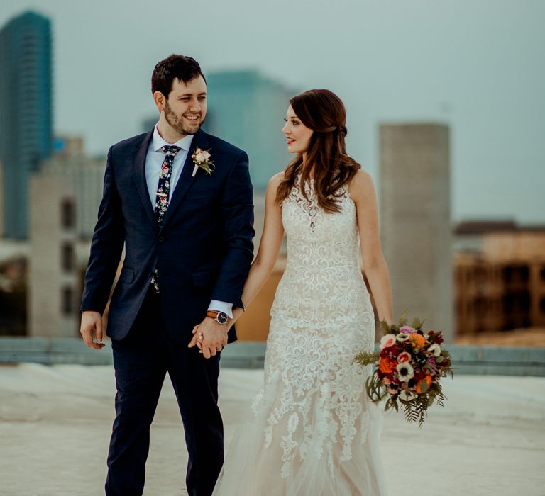 Bride in Sleeveless High-Neck Stella York Gown with Lace Overlay and Gold Underskirt | Half Up Half Down Wedding Hair | Bride Wearing Red Lipstick | Groom in Navy Suit and Floral Tie | Pink Buttonhole | Colourful Bridal Bouquet of Pink, Orange and White Flowers with Ferns, Eucalyptus and Greenery | Rifle Paper Co. Trainers for Informal Wedding in Fort Worth, Texas | Paul &amp; Nanda Photography