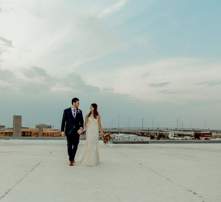 Bride in Sleeveless High-Neck Stella York Gown with Lace Overlay and Gold Underskirt | Half Up Half Down Wedding Hair | Bride Wearing Red Lipstick | Groom in Navy Suit and Floral Tie | Pink Buttonhole | Colourful Bridal Bouquet of Pink, Orange and White Flowers with Ferns, Eucalyptus and Greenery | Rifle Paper Co. Trainers for Informal Wedding in Fort Worth, Texas | Paul &amp; Nanda Photography