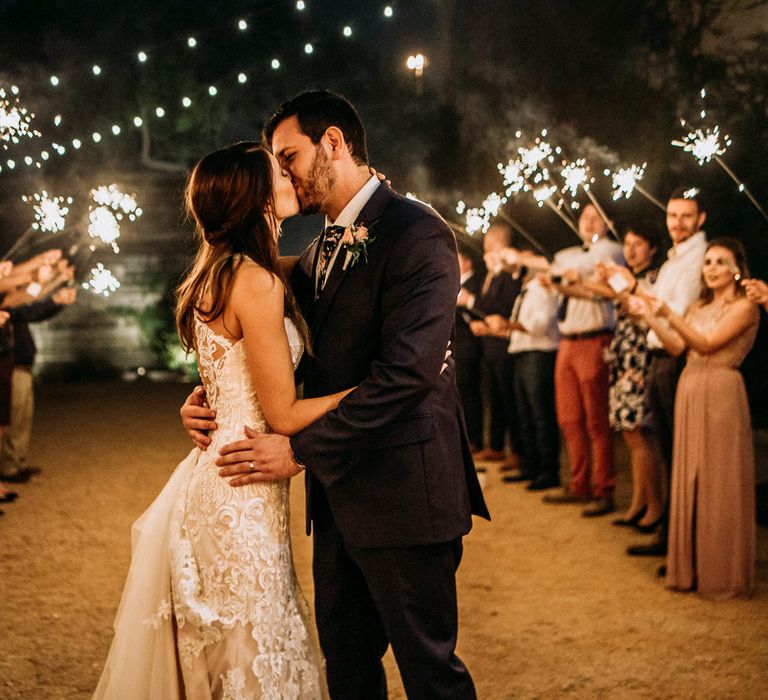 Sparkler Send Off | Bride in Sleeveless High-Neck Stella York Gown with Lace Overlay and Gold Underskirt | Groom in Navy Suit and Floral Tie | Pink Buttonhole | Festoon Lights | Rifle Paper Co. Trainers for Informal Wedding in Fort Worth, Texas | Paul &amp; Nanda Photography