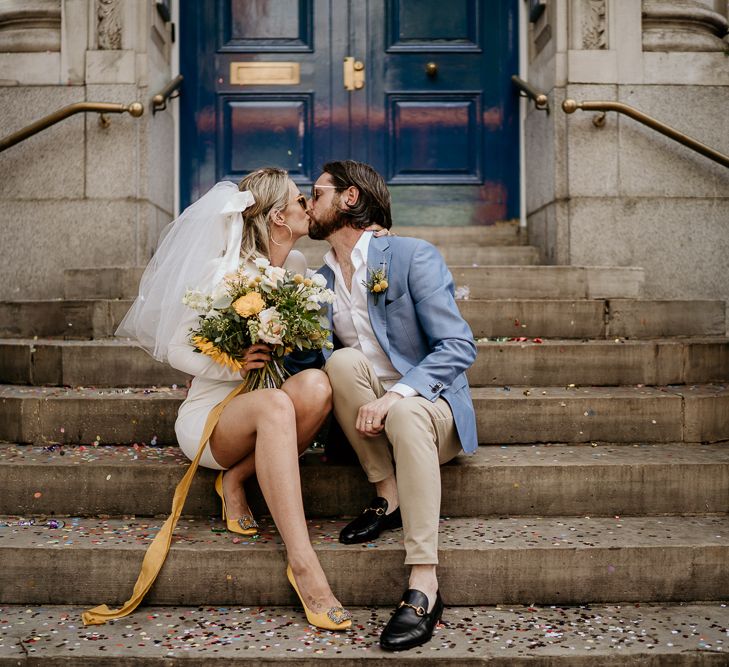 Stylish bride in short wedding dress with yellow shoes and bouquet and groom in blue blazer at COVID wedding