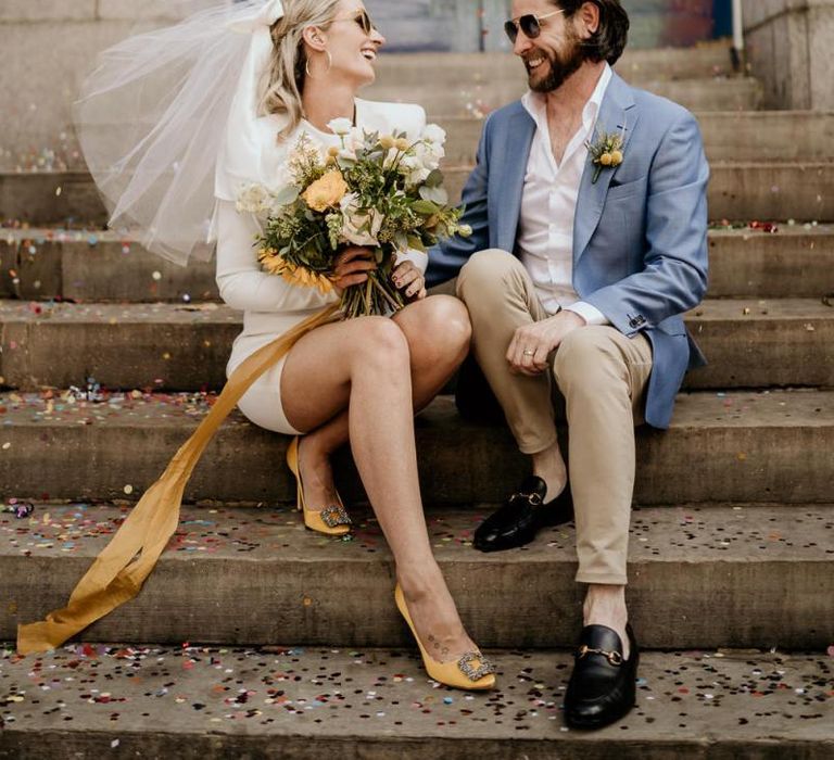 Stylish bride in short wedding dress with yellow shoes and bouquet and groom in blue blazer and chinos sitting on a step