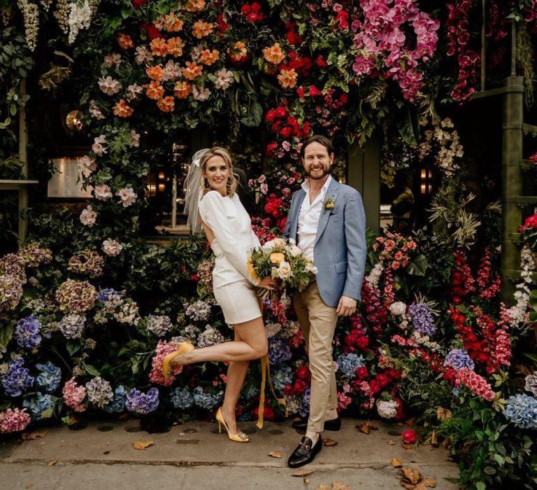 Stylish bride in short wedding dress and groom in blue blazer standing in front of a flower installation