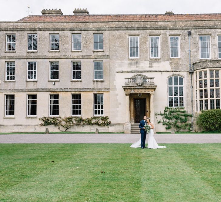 Bride in Fitted Mikaella Wedding Dress with Flower Crown and Groom in Bespoke Navy Check Suit by Territo Standing in Front of Elmore Court