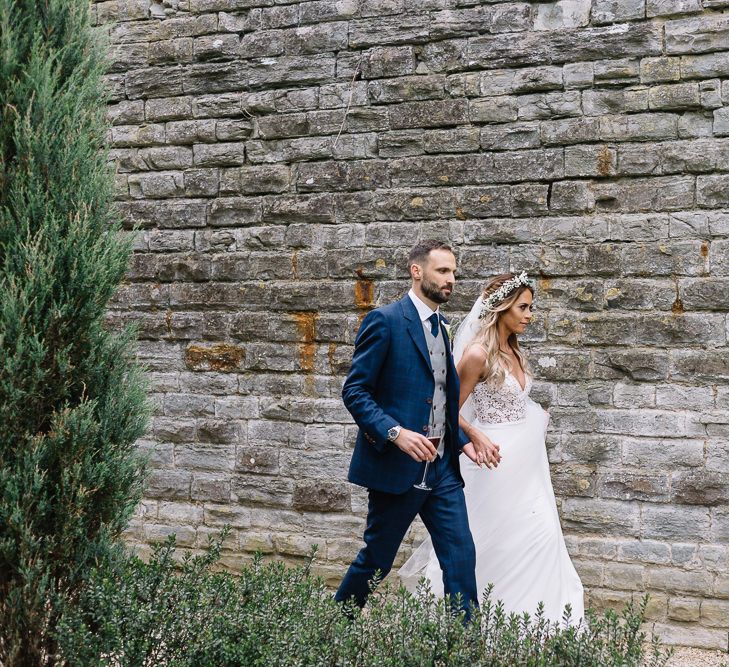 Bride in Fitted Mikaella Wedding Dress with Flower Crown and Groom in Bespoke Navy Check Suit by Territo Walking in the Grounds of Elmore Court