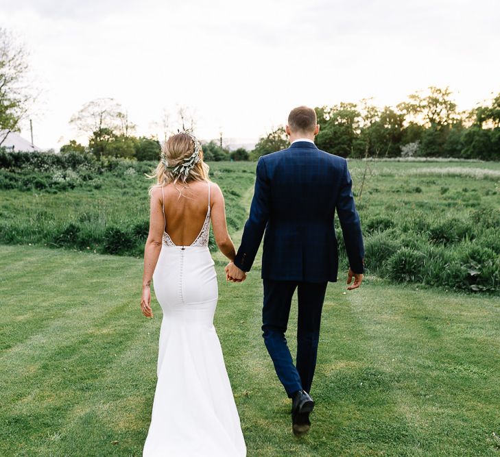 Bride in Fitted Mikaella Wedding Dress with Low Back and Flower Crown and Groom in Bespoke Navy Check Suit Holding Hands