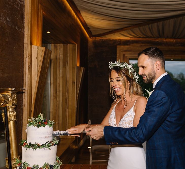 Bride in Fitted Mikaella Wedding Dress  and Flower Crown and Groom in Bespoke Navy Check Suit Cutting the Wedding Cake