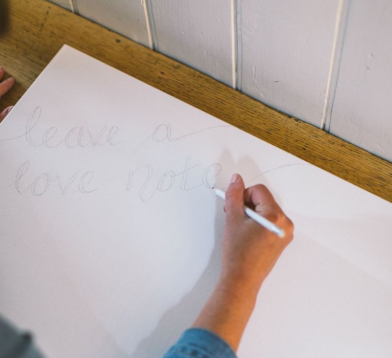 Person writing the words 'Leave a love note' onto a box canvas in pencil