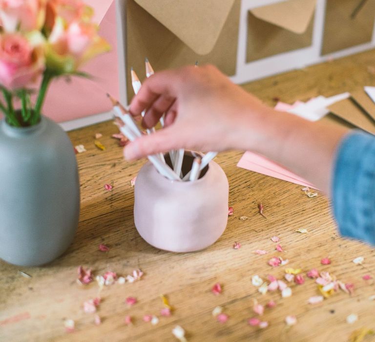 Person putting white pencils into a dusky pink pot next to a grey vase filled with pale pink antique roses as part of a DIY wedding card table set up