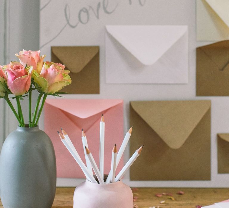 Pink ceramic pot filled with white pencils and muted grey vase filled with pale pink roses as part of a DIY wedding card table