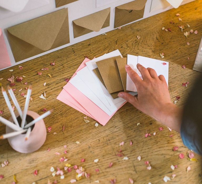 Person laying notecards on top of a wedding card table, next to a pot of pencils and a box for guests to put wedding cards inside