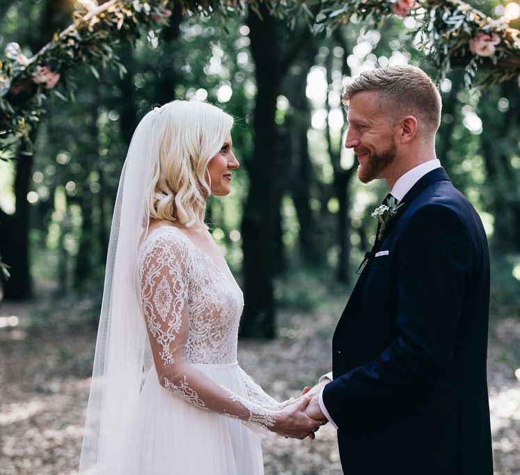 Outdoor Wedding Service | Bride in Long Sleeved Anna Kara Wedding Dress with Lace Bodice and Soft Skirt | Britten Weddings Floor Length Veil | Groom in Dark Navy Hugo Boss Suit with Waistcoat and Black Patent Shoes by Common Projects | Fairy Light Moon Gate Altar | Puglian Countryside Wedding with Fairy Light Altar and Olive Grove Aperitivo | Figtree Wedding Photography