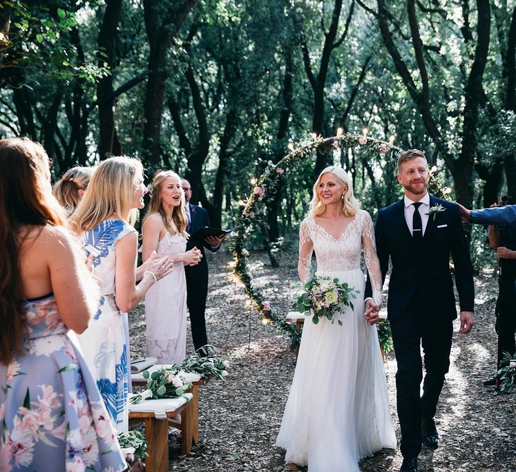 Just Married | Bride in Long Sleeved Anna Kara Wedding Dress with Lace Bodice and Soft Skirt | Britten Weddings Floor Length Veil | Bridal Bouquet of White, Soft Pink, Purple and Yellow Flowers with Greenery | Groom in Dark Navy Hugo Boss Suit with Waistcoat and Black Patent Shoes by Common Projects | Fairy Light Moon Gate Altar | Puglian Countryside Wedding with Fairy Light Altar and Olive Grove Aperitivo | Figtree Wedding Photography