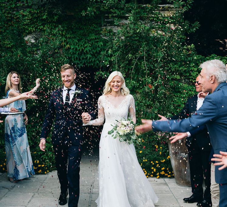 Confetti Throw | Bride in Long Sleeved Anna Kara Wedding Dress with Lace Bodice and Soft Skirt | Britten Weddings Floor Length Veil | Bridal Bouquet of White, Soft Pink, Purple and Yellow Flowers with Greenery | Groom in Dark Navy Hugo Boss Suit with Waistcoat and Black Patent Shoes by Common Projects | Puglian Countryside Wedding with Fairy Light Altar and Olive Grove Aperitivo | Figtree Wedding Photography