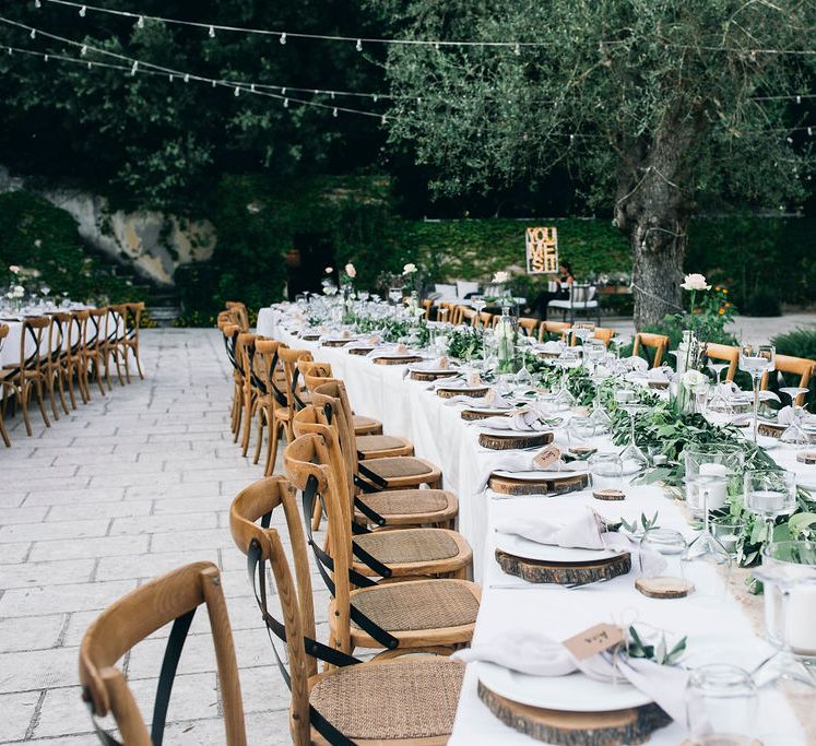 Outdoor Wedding Reception Decor at Tenuta Tresca | Long Tables Snaking Through Courtyard | Foliage Table Runners | Festoon Lights | Knotted Lilac Napkins | Wood Slice Place Mats | Tall Stemmed Candle Holders | Puglian Countryside Wedding with Fairy Light Altar and Olive Grove Aperitivo | Figtree Wedding Photography