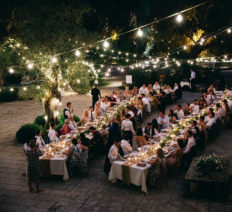 Outdoor Wedding Breakfast at Tenuta Tresca | Long Tables Snaking Through Courtyard | Foliage Table Runners | Festoon Lights | Tall Stemmed Candle Holders | Fairy Light Moon Gate | Puglian Countryside Wedding with Fairy Light Altar and Olive Grove Aperitivo | Figtree Wedding Photography