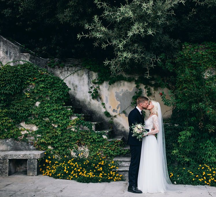 Bride in Long Sleeved Anna Kara Wedding Dress with Lace Bodice and Soft Skirt | Britten Weddings Floor Length Veil | Bridal Bouquet of White, Soft Pink, Purple and Yellow Flowers with Greenery | Groom in Dark Navy Hugo Boss Suit with Waistcoat and Black Patent Shoes by Common Projects | Puglian Countryside Wedding with Fairy Light Altar and Olive Grove Aperitivo | Figtree Wedding Photography