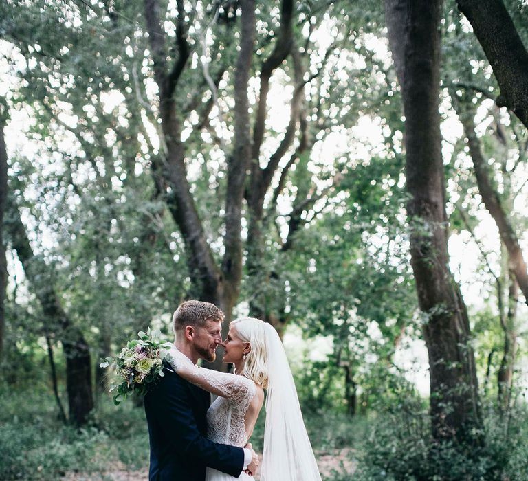 Bride in Long Sleeved Anna Kara Wedding Dress with Lace Bodice and Soft Skirt | Britten Weddings Floor Length Veil | Bridal Bouquet of White, Soft Pink, Purple and Yellow Flowers with Greenery | Groom in Dark Navy Hugo Boss Suit with Waistcoat and Black Patent Shoes by Common Projects | Puglian Countryside Wedding with Fairy Light Altar and Olive Grove Aperitivo | Figtree Wedding Photography