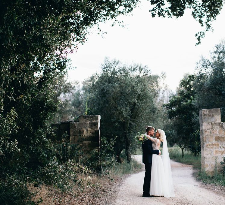 Bride in Long Sleeved Anna Kara Wedding Dress with Lace Bodice and Soft Skirt | Britten Weddings Floor Length Veil | Bridal Bouquet of White, Soft Pink, Purple and Yellow Flowers with Greenery | Groom in Dark Navy Hugo Boss Suit with Waistcoat and Black Patent Shoes by Common Projects | Puglian Countryside Wedding with Fairy Light Altar and Olive Grove Aperitivo | Figtree Wedding Photography