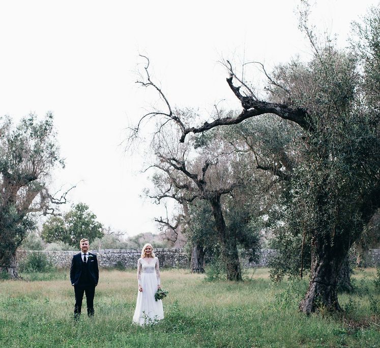 Bride in Long Sleeved Anna Kara Wedding Dress with Lace Bodice and Soft Skirt | Britten Weddings Floor Length Veil | Bridal Bouquet of White, Soft Pink, Purple and Yellow Flowers with Greenery | Groom in Dark Navy Hugo Boss Suit with Waistcoat and Black Patent Shoes by Common Projects | Puglian Countryside Wedding with Fairy Light Altar and Olive Grove Aperitivo | Figtree Wedding Photography