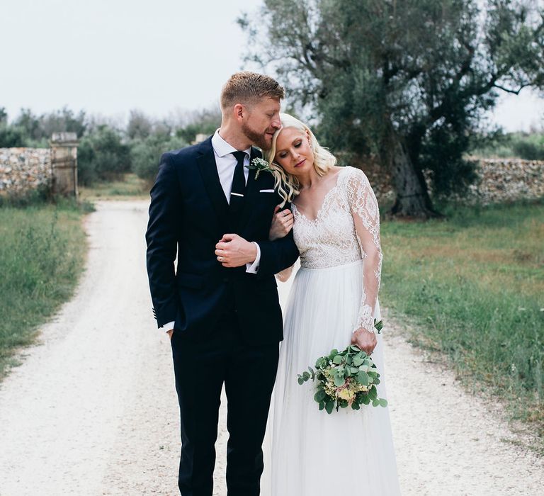 Bride in Long Sleeved Anna Kara Wedding Dress with Lace Bodice and Soft Skirt | Britten Weddings Floor Length Veil | Bridal Bouquet of White, Soft Pink, Purple and Yellow Flowers with Greenery | Groom in Dark Navy Hugo Boss Suit with Waistcoat and Black Patent Shoes by Common Projects | Puglian Countryside Wedding with Fairy Light Altar and Olive Grove Aperitivo | Figtree Wedding Photography