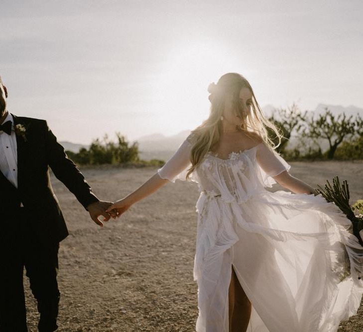 Bride in Ruffles Wedding Dress and Groom in Black Tuxedo Holding Hands During Golden Hour