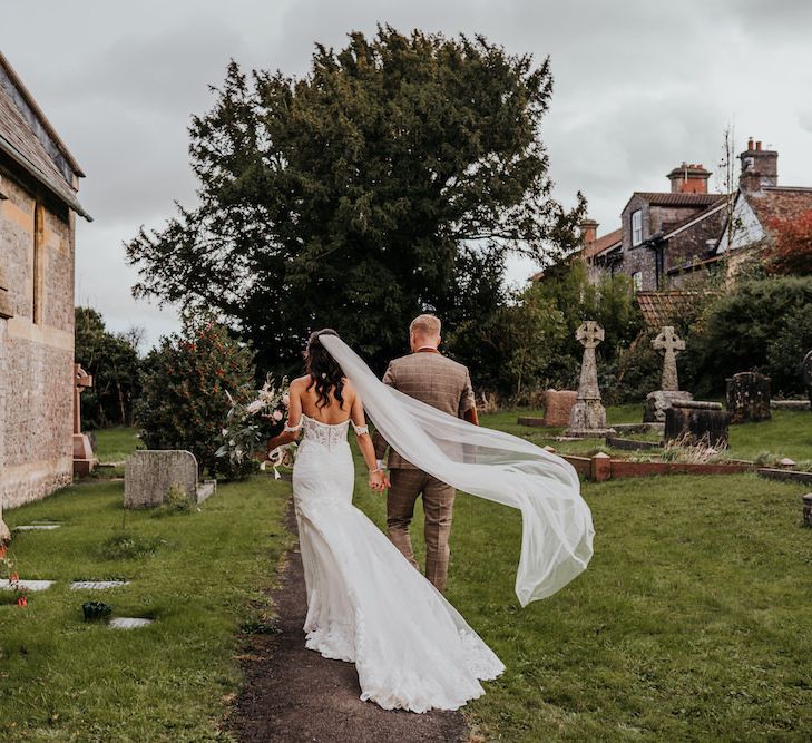 Bride in lace Martina Liana wedding dress with billowing veil walking through the church courtyard