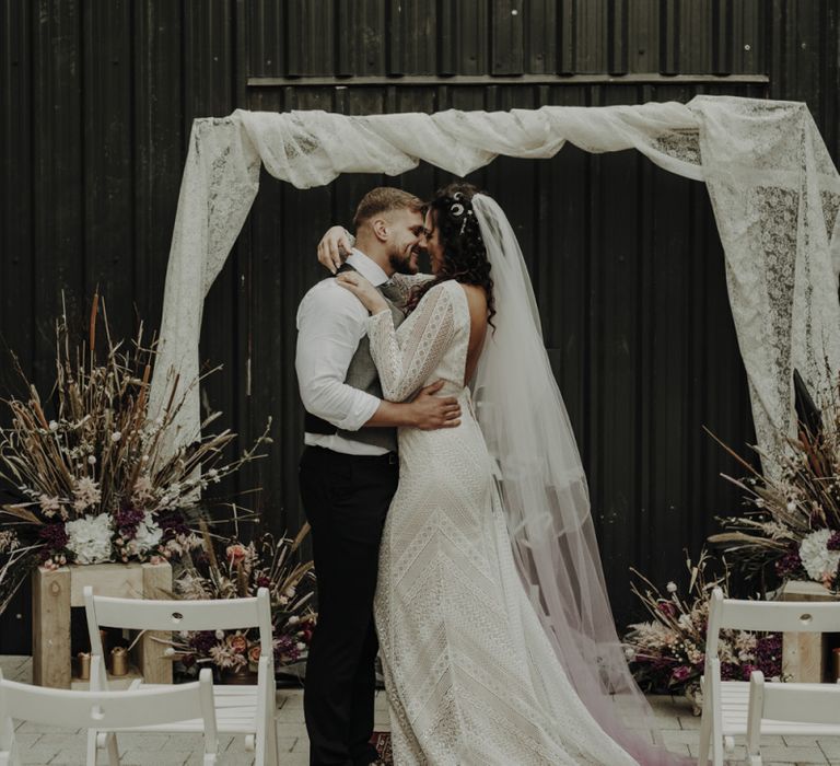 Boho Bride in Lace Wedding Dress and Groom Kissing at the Altar with Drapes, Woven Rug, Bold Sign and Dried Flower Arrangements