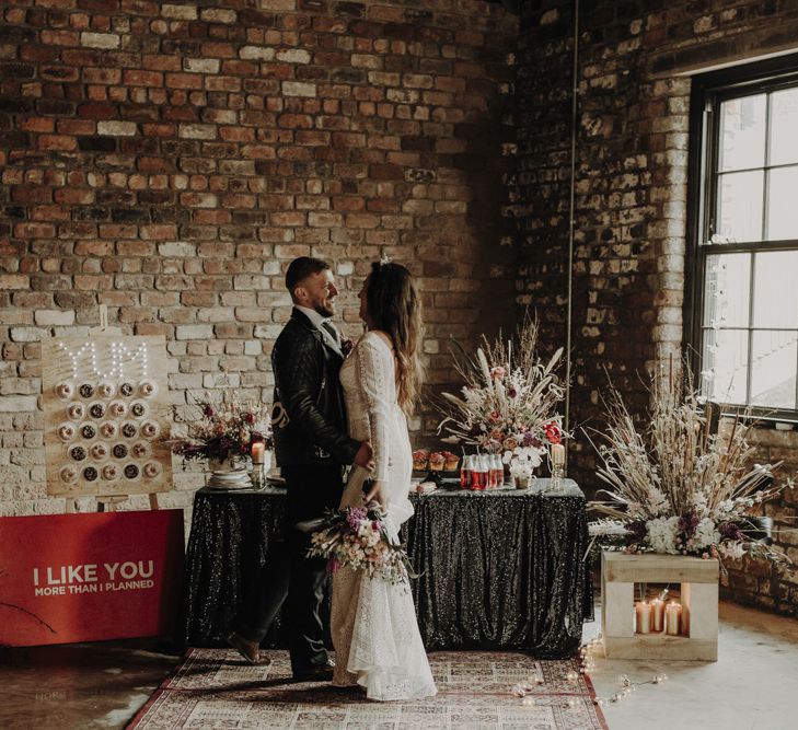 Boho Bride in Lace Wedding Dress and Groom in Leather Jacket Standing at Dessert Table with Doughnut Wall, Wedding Cakes and Dried Flower Arrangements