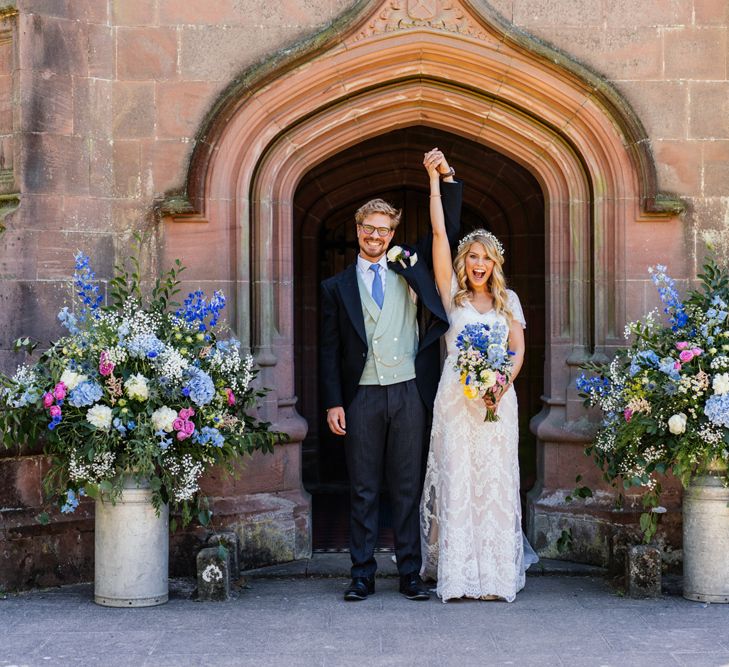 Bride in Lace Essence of Australia Wedding Dress and Groom in Morning Suit Exiting the Church with Pink and Blue Wildflower Filled Milk Churns