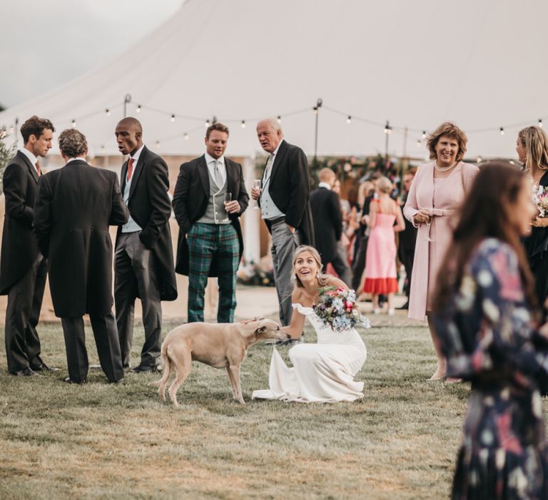 Bride in off the shoulder wedding dress stroking her pet dog during outdoor reception