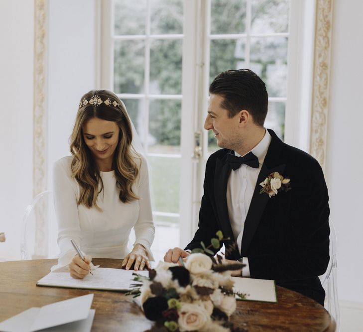 Bride in Emma Beaumont Wedding Dress and Tilly Thomas Lux Headdress and Groom in Black Tie Suit Signing the Register